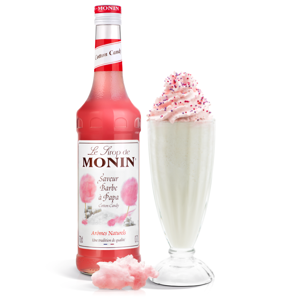 A bottle of Monin cotton candy syrup stands next to a tall glass filled with a white milkshake topped with pink cotton candy syrup and sprinkles, with loose pink cotton candy in front on a white background.