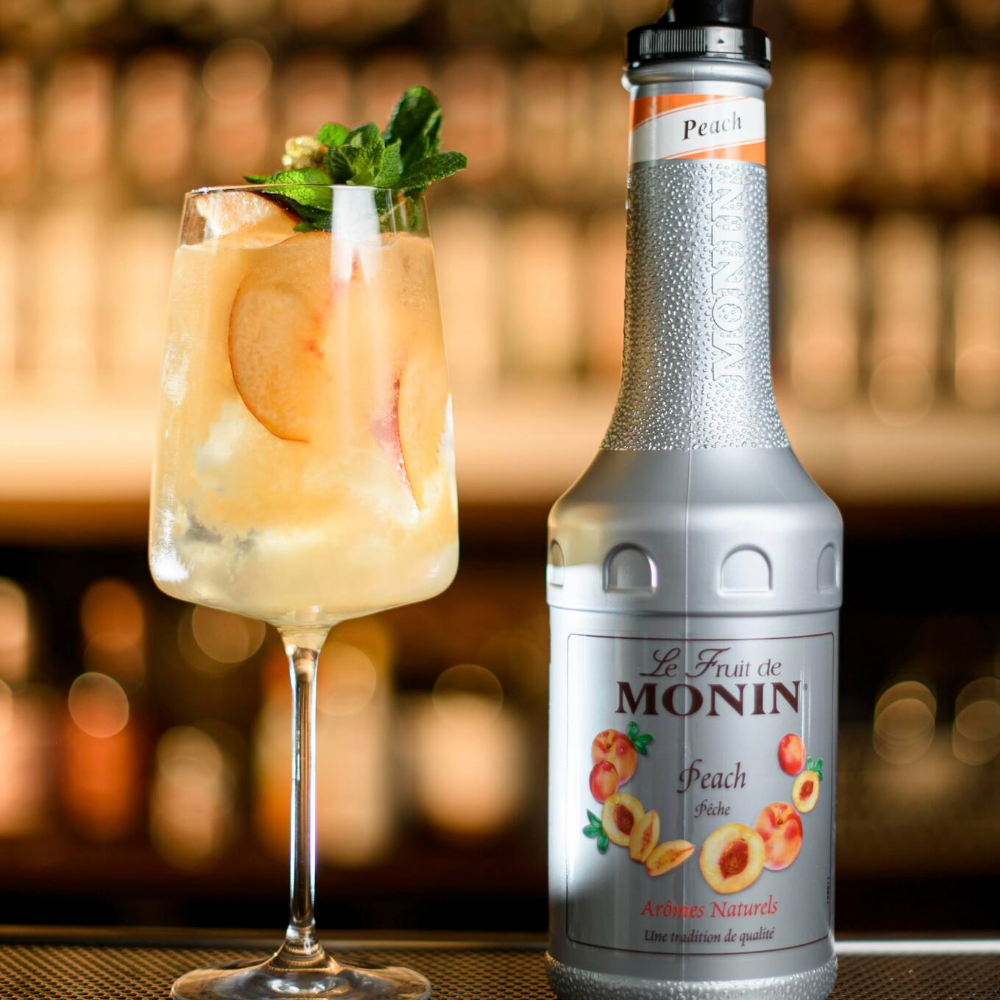 A bottle of Le Fruit de Monin Peach Syrup stands next to a prepared peach-colored cocktail in a highball glass on a bar counter, with a blurred bar background.
