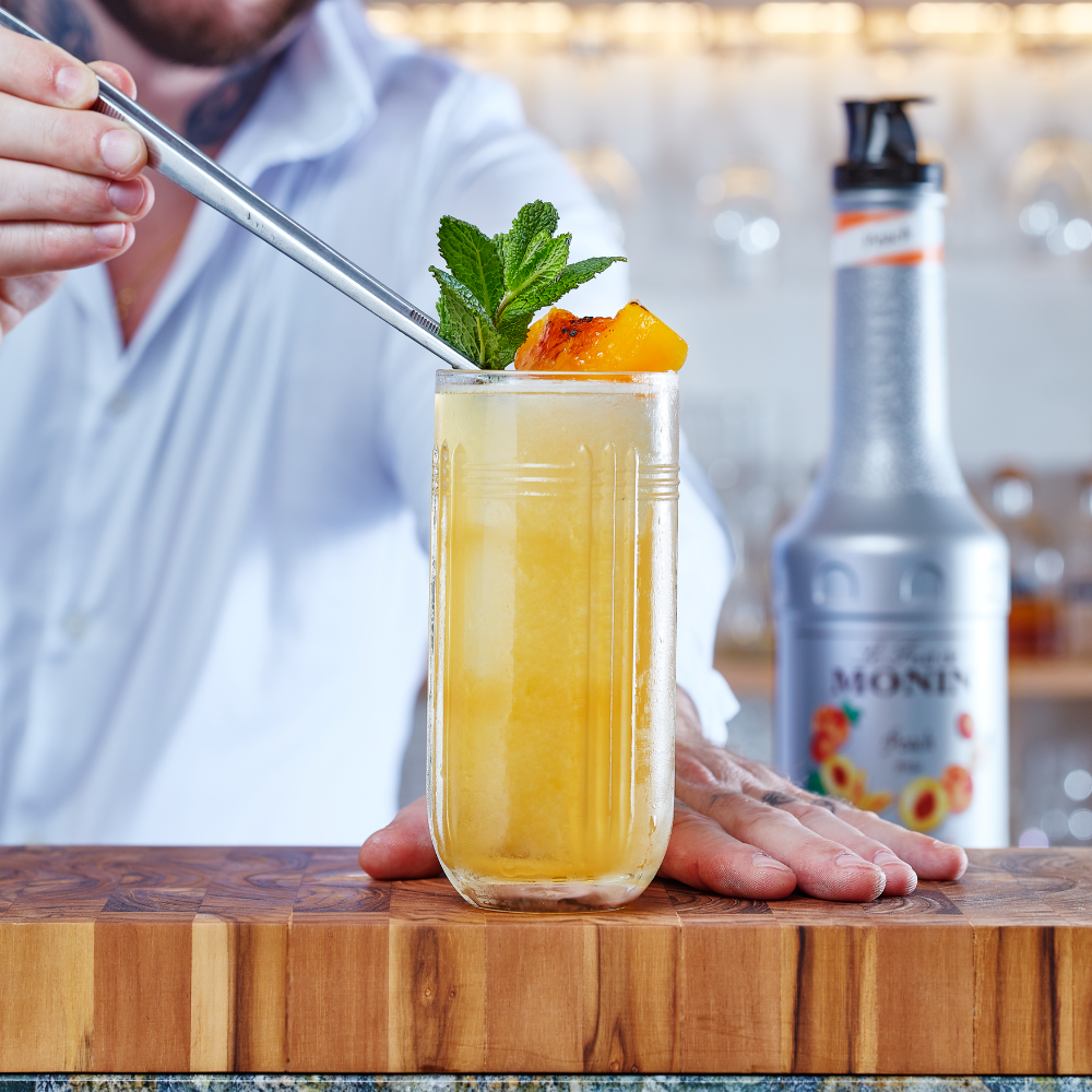 A bartender in a white shirt holds a metal straw over a tall glass of yellow-orange cocktail garnished with mint and a peach slice, set on a wooden bar counter with a Monin bottle blurred in the background.