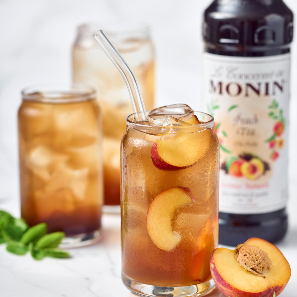 Monin Peach Tea Syrup 1ltr Glass of iced tea with peach slices, Monin Peach Tea Syrup bottle in the background - Discount Coffee