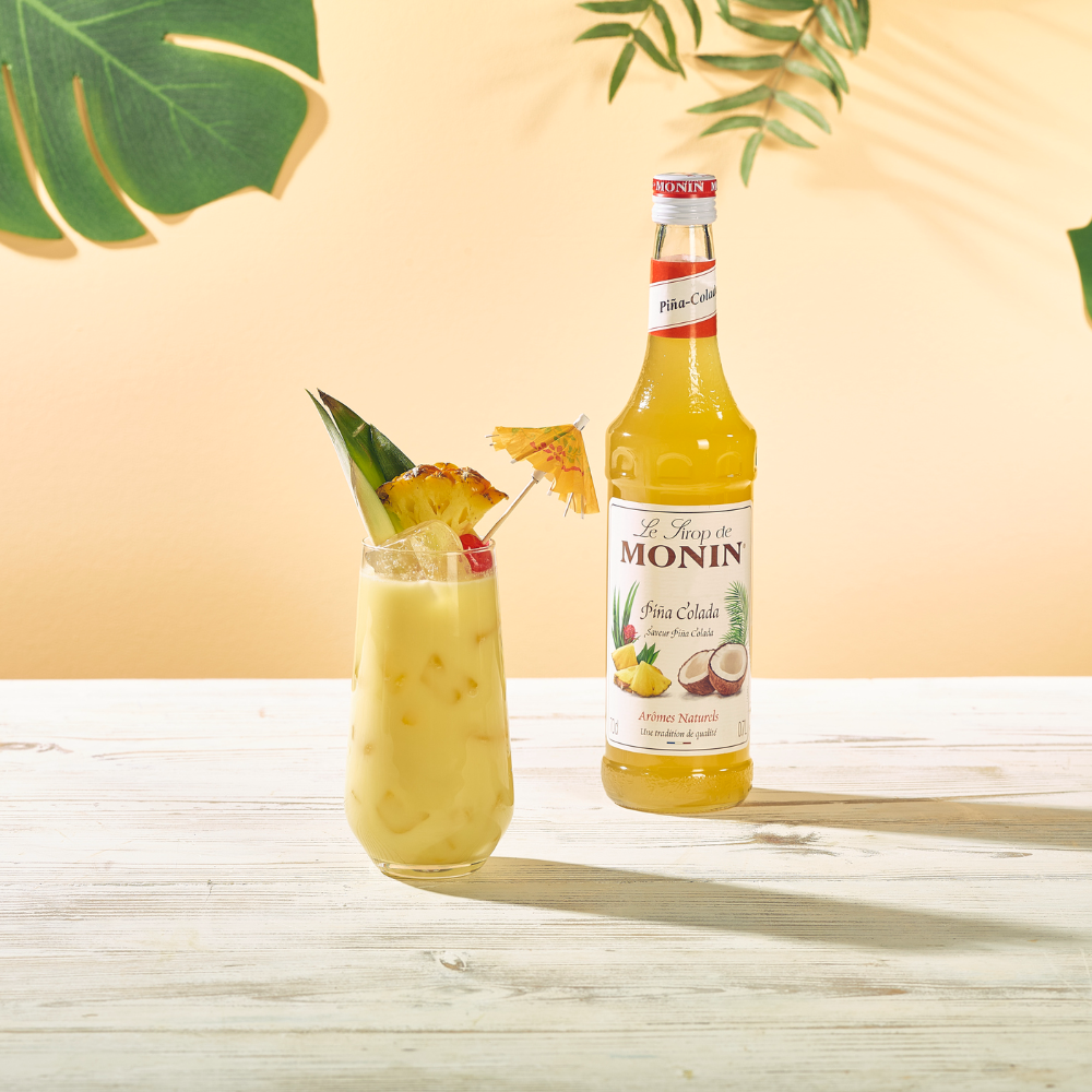 Monin Pina Colada Syrup Bottle 70 cl. A picture of a Pina Colada cocktail drink in a tall glass made with 23% natural juice next to a Monin bottle of Pina Colada syrup on a tropical themed background - Discount Coffee
