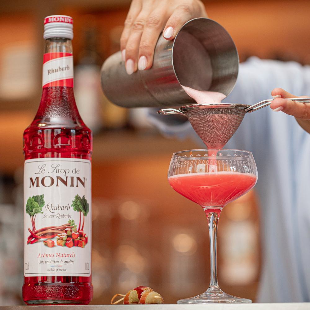 Monin Rhubarb Flavouring Syrup (700ml) A Picture of a Person Pouring a Rhubarb Cocktail From a Cocktail Shaker Into a Cocktail Glass - Discount Coffee