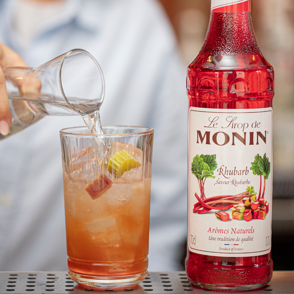 Monin Rhubarb Syrup Bottle (700ml) Pouring a Bottle of Cold Water into a Tall Glass of Iced Rhubarb Soda - Discount Coffee