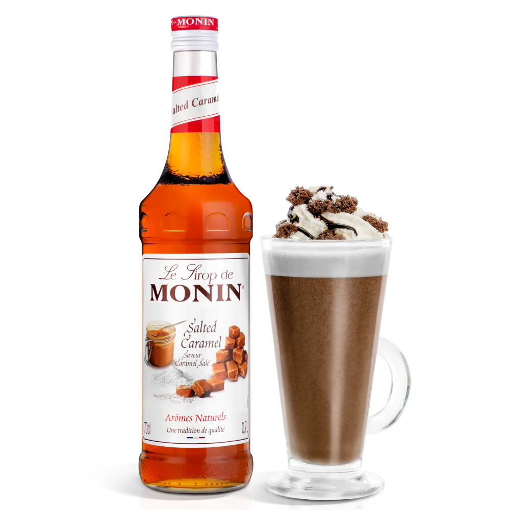 A bottle of Monin Salted Caramel syrup stands next to a tall glass of hot chocolate topped with whipped cream, chocolate drizzle, and a sprinkle of caramel pieces, on a white background.