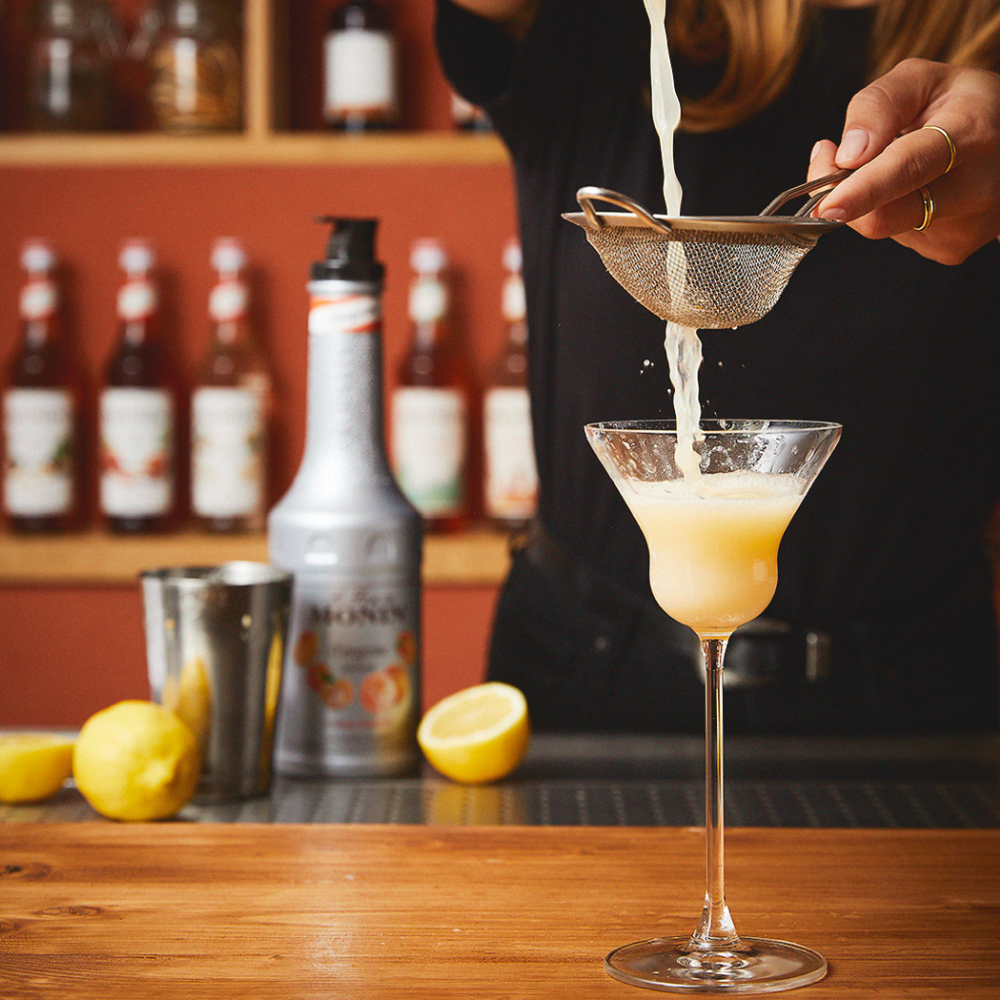 A bartender strains a pale yellow drink into a martini glass using a metal strainer, with lemons, bottles, and a shaker in a warm bar setting.
