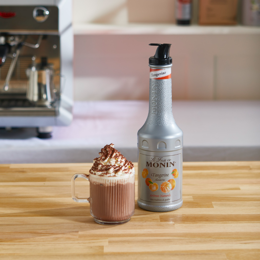 A bottle of Monin Tangrite puree stands on a wooden counter beside a glass mug filled with a hot chocolate or coffee drink topped with whipped cream and chocolate shavings, with a blurred kitchen or bar background.