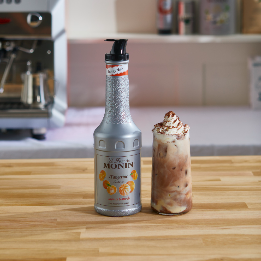 A bottle of Monin Tangine puree 1 litre with a silver body and black pump stands on a wooden counter next to a tall glass filled with an iced coffee or latte topped with whipped cream and cocoa dusting.