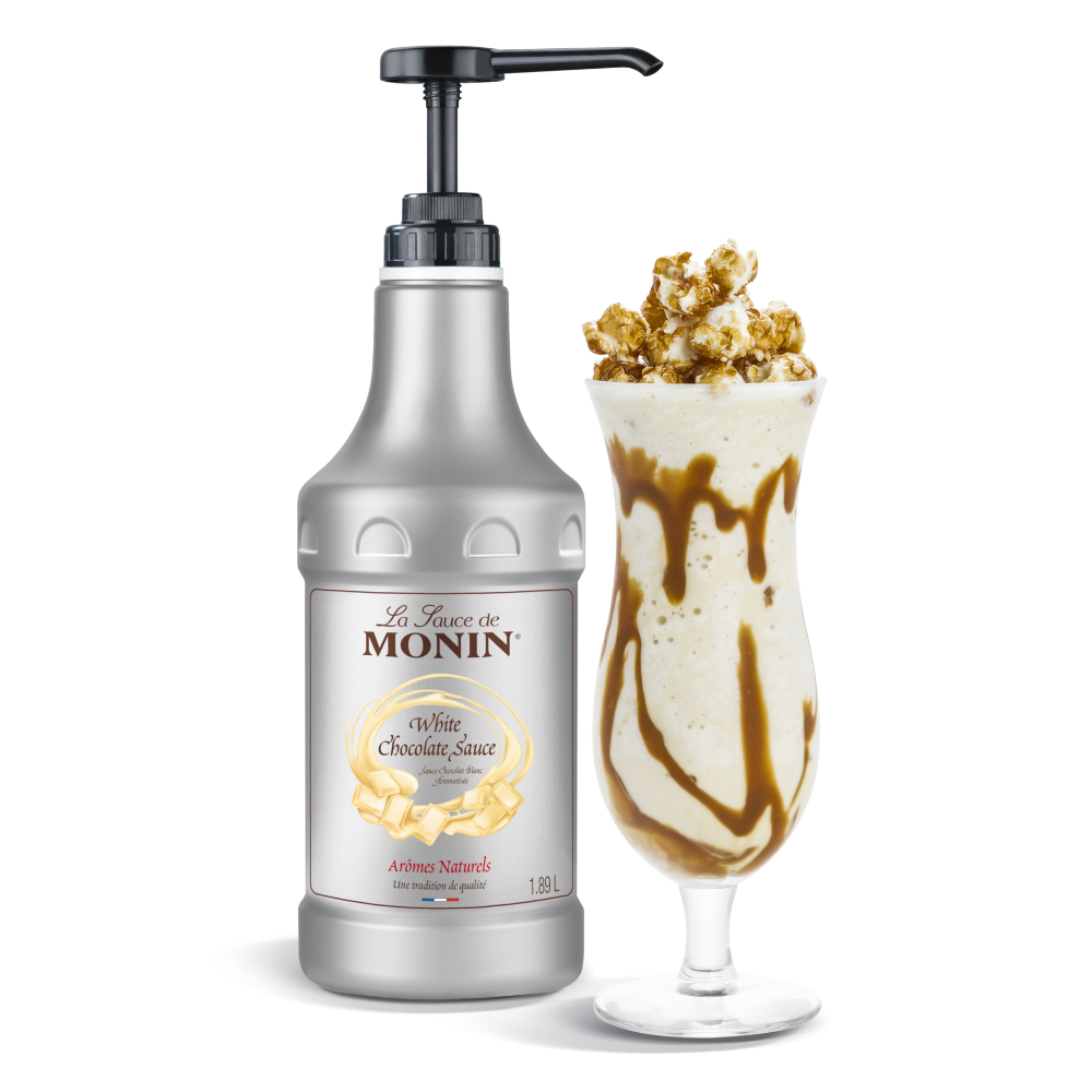 A bottle of 1.89L Monin White Chocolate Sauce with a black pump next to a tall glass of milkshake topped with caramel sauce and caramel popcorn, on a white background.